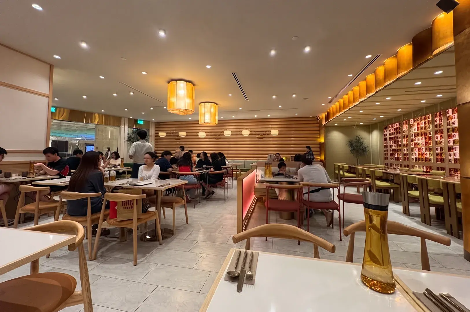 Warmly lit, open-plan restaurant dining room with light wooden furniture and glowing drum lamps.