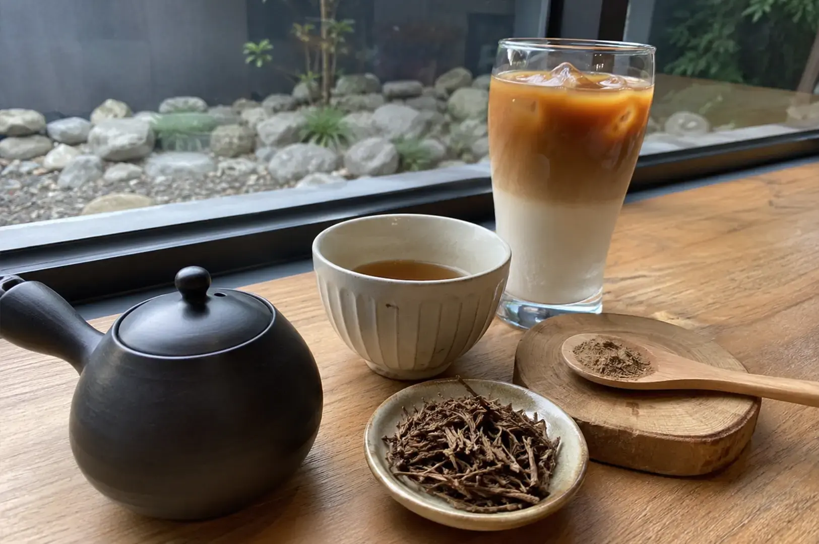 A dark ceramic teapot sits beside a small cup of brewed tea and a glass of iced, layered latte on a wooden table, accompanied by a small dish of dried tea stalks and a wooden spoon holding a mound of ground tea powder.