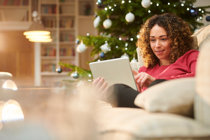 Young woman laying on the sofa in front of a Christmas tree using a tablet. 