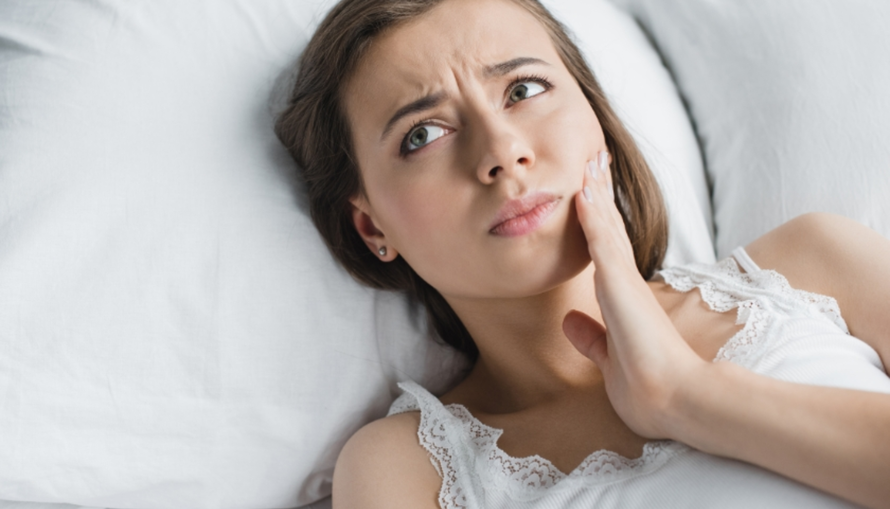 Kvinne ligger i sengen med hånden på kinnet og uttrykk av smerte.