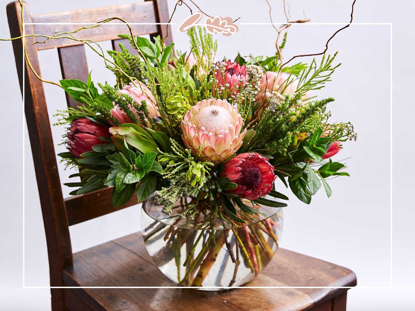 A stunning arrangement of proteas and greenery in a clear glass bowl on a wooden chair - Fabulous Flowers and Gifts