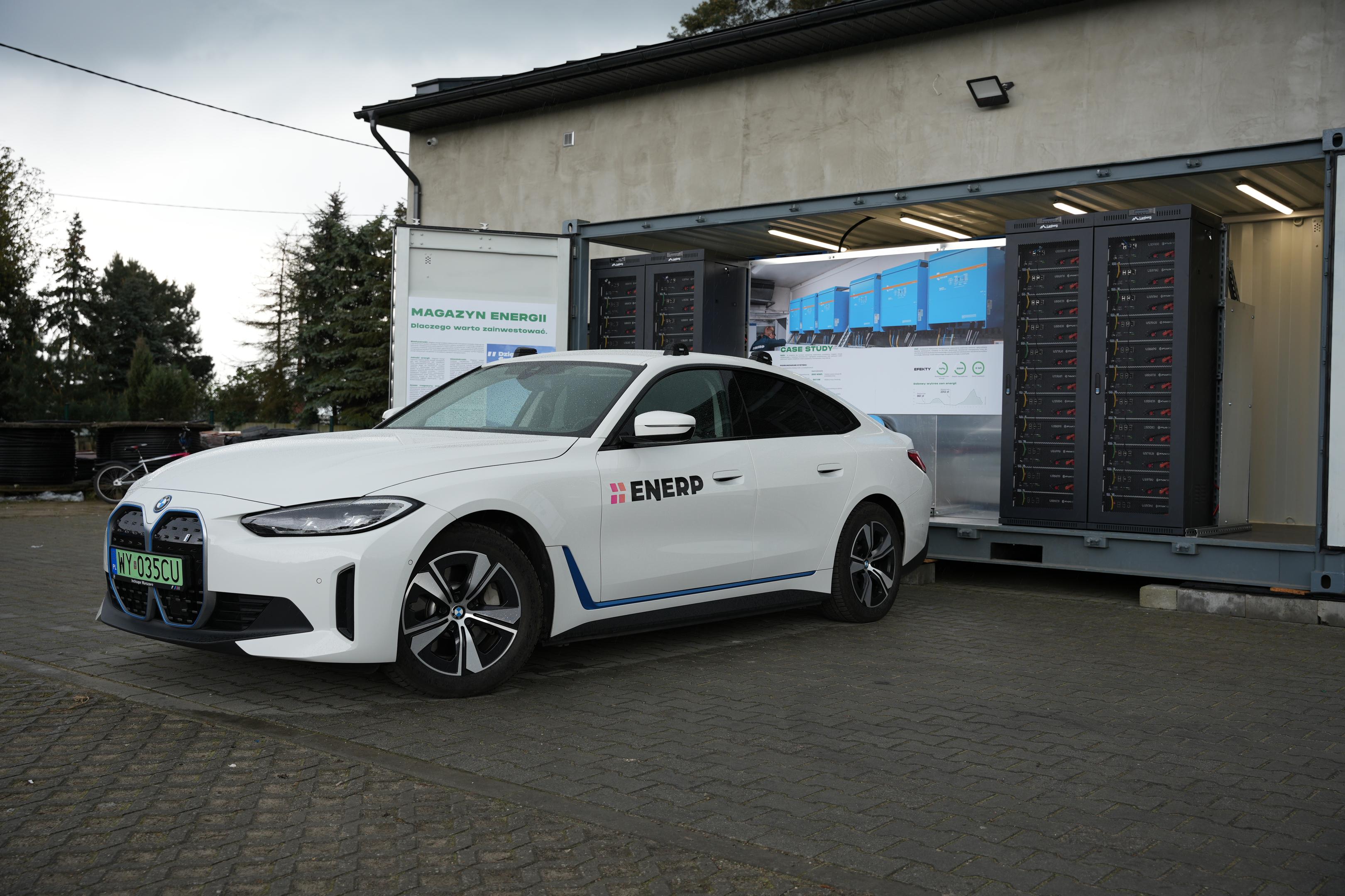Na dachu jednorodzinnego domu znajdują się panele fotowoltaiczne, a na podjeździe stoi samochód elektryczny, symbolizujący nowoczesne podejście do energii i niższe koszty eksploatacji samochodu elektrycznego. Obrazek ukazuje harmonię między ekologicznym stylem życia a nowoczesnym transportem.