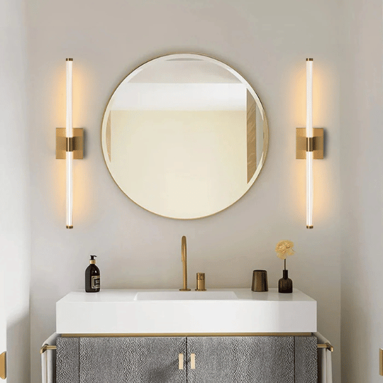 Stylish bathroom interior with a round wall-mounted mirror framed in gold, flanked by two vertical wall sconces emitting warm light. Below, a modern vanity with a white countertop, gold faucet, and textured gray cabinet doors holds decorative items including a small vase with a single flower and a soap dispenser.