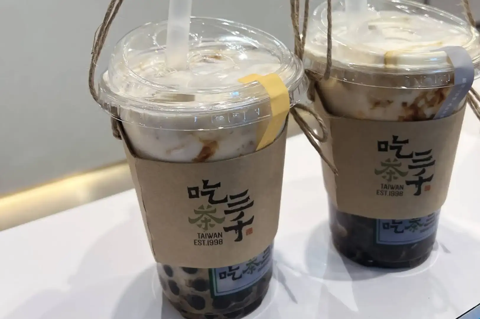 Two iced CHICHA San Chen milk tea beverages with black tapioca pearls rest on a white table, fitted with kraft paper sleeves displaying the brand's Chinese calligraphy logo and suspended by traditional rustic hemp rope carrying handles.