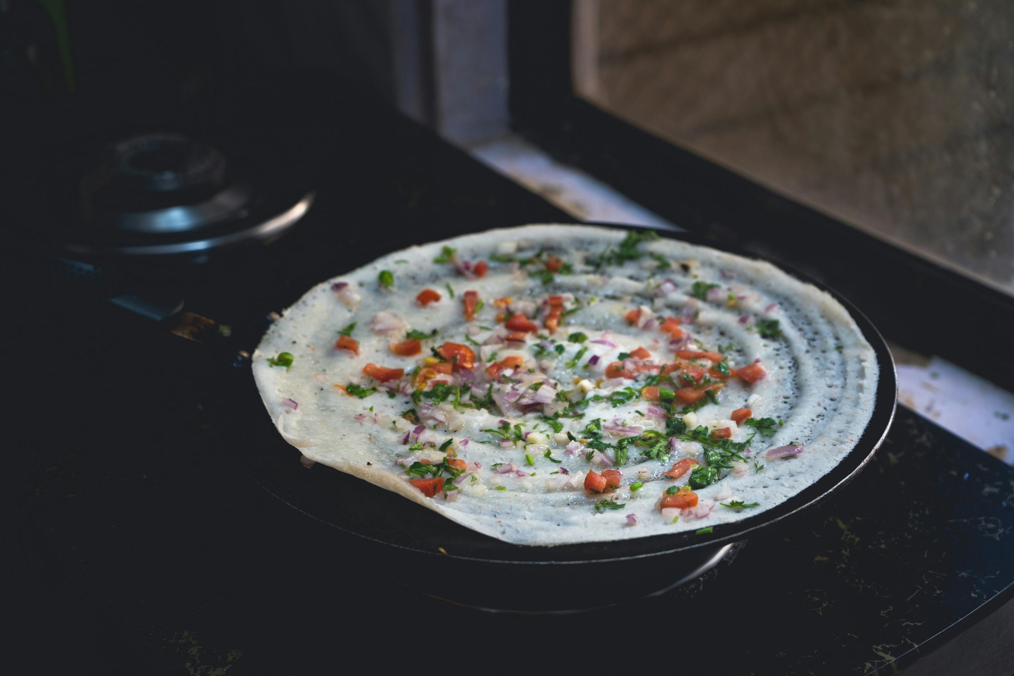  A thin, circular dosa cooks on a flat black griddle, its surface speckled with a vibrant mix of chopped red tomatoes, purple onions, and fresh green herbs. The pan sits atop a dark stovetop near a window, capturing the simple beauty of a fresh meal being prepared in a home kitchen.