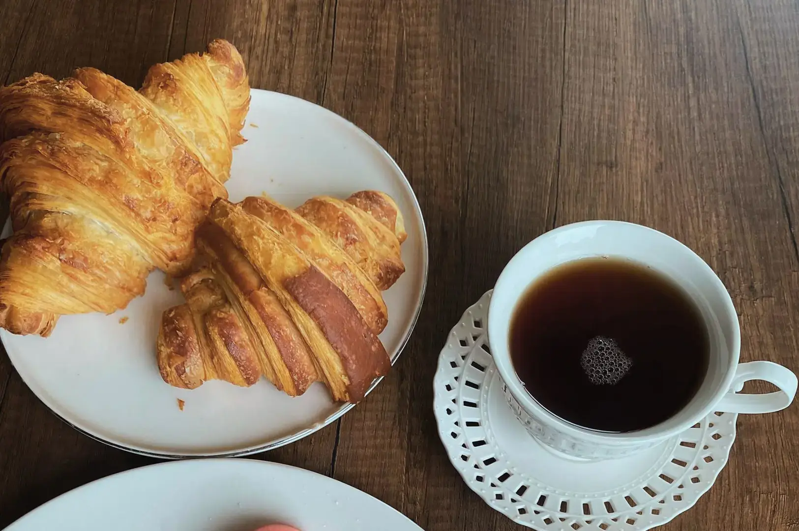 An overhead shot shows a breakfast setup resting on a rustic wooden table surface. On the left, two flaky, golden-brown croissants are served on a simple white plate. To the right sits a delicate white ceramic teacup filled with dark tea, resting on a matching saucer that features an intricate, open-lace geometric cutout pattern around its rim.
