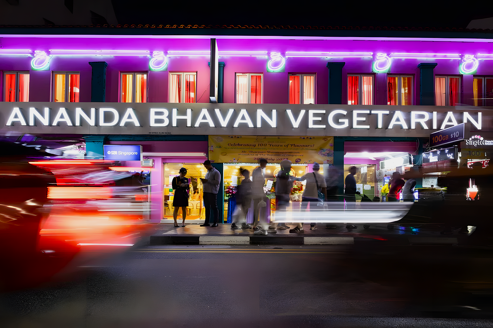 Nighttime scene of a brightly lit vegetarian restaurant with neon pink and blue lights. Blurred motion of cars and people creates a lively, vibrant atmosphere.