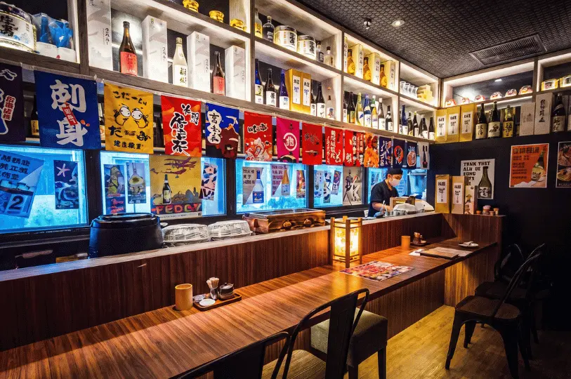 Cozy, vibrant interior of a Japanese izakaya in Singapore featuring a long wooden bar counter, rows of sake bottles on shelves, and colorful Japanese banners hanging against the wall.