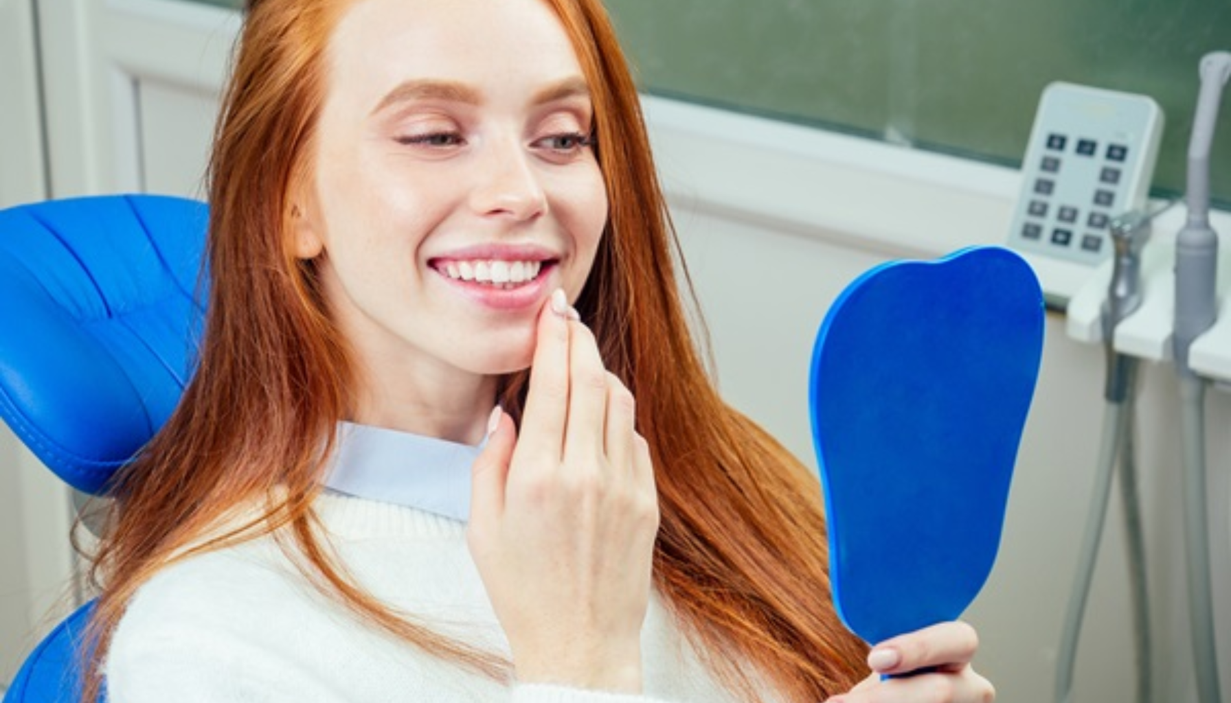 "Rødhåret kvinne i tannlegestol smiler og holder speil mens hun ser på tennene sine."