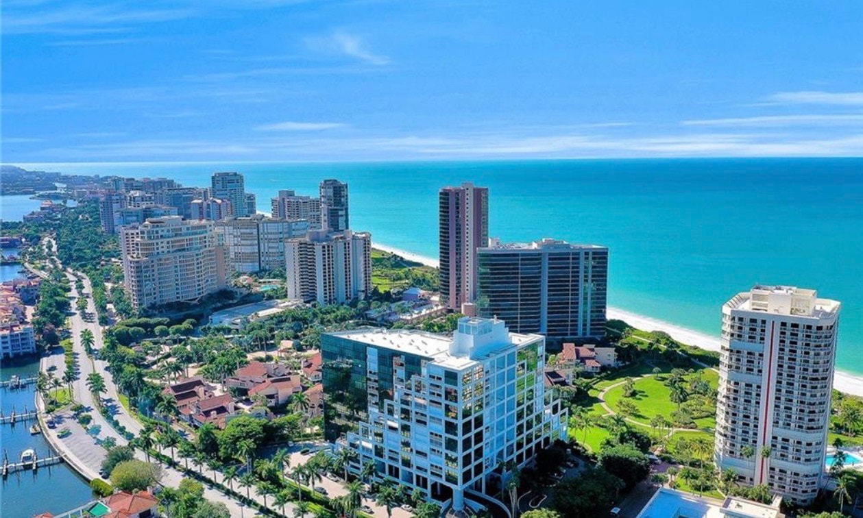 An aerial view of Park Shore in Naples, Florida, showcases the stunning Gulf coastline and the Venetian Bay waterfront community, highlighting luxury real estate options like mid-rise condos and single-family homes. The image captures the picturesque views of private beaches, waterfront properties, and the vibrant lifestyle of Park Shore residents, including access to upscale shopping and dining at the nearby Village Shops.