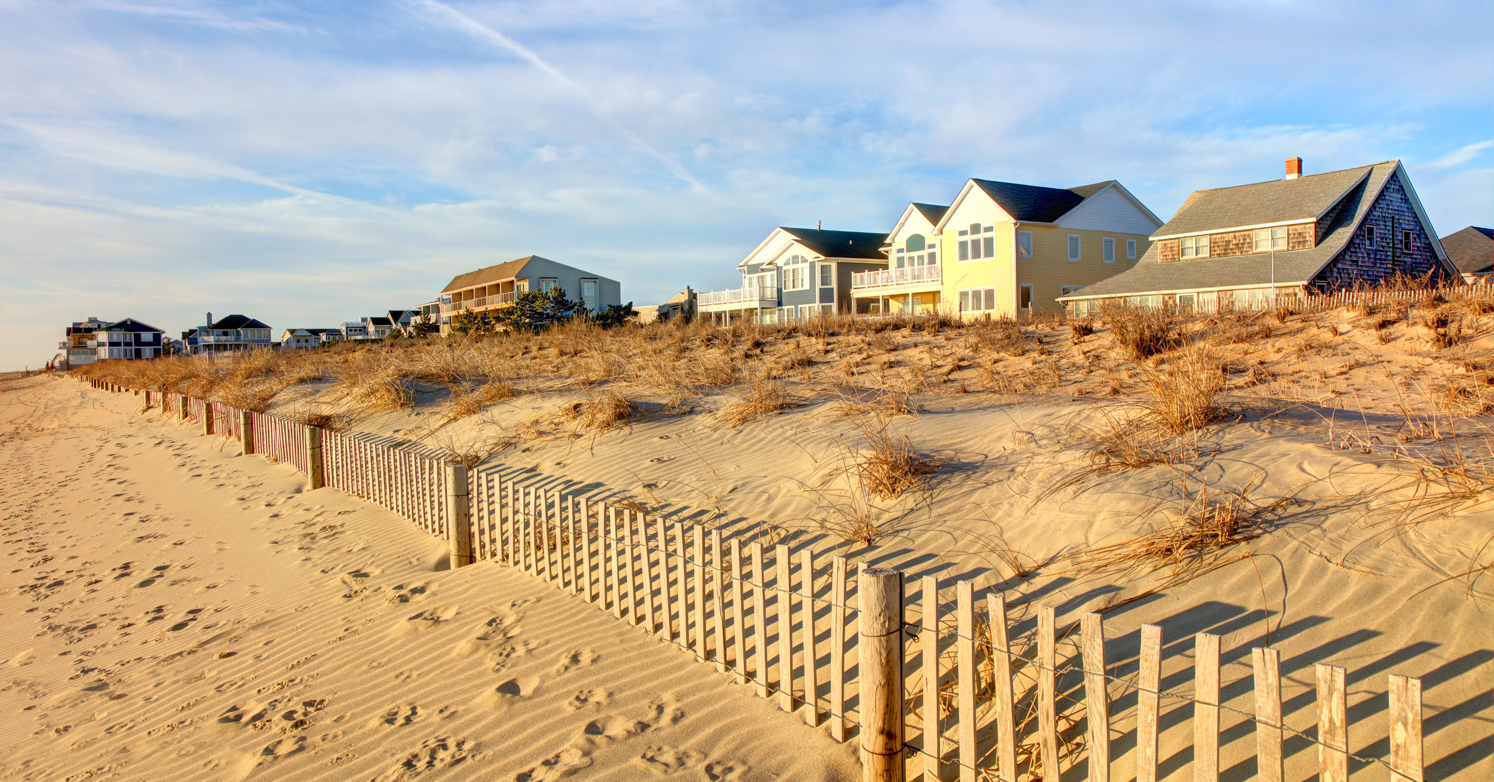 Beachfront vacation homes in North Cape May, NJ, overlooking sand dunes and a wooden fence along a quiet bay beach at sunset.