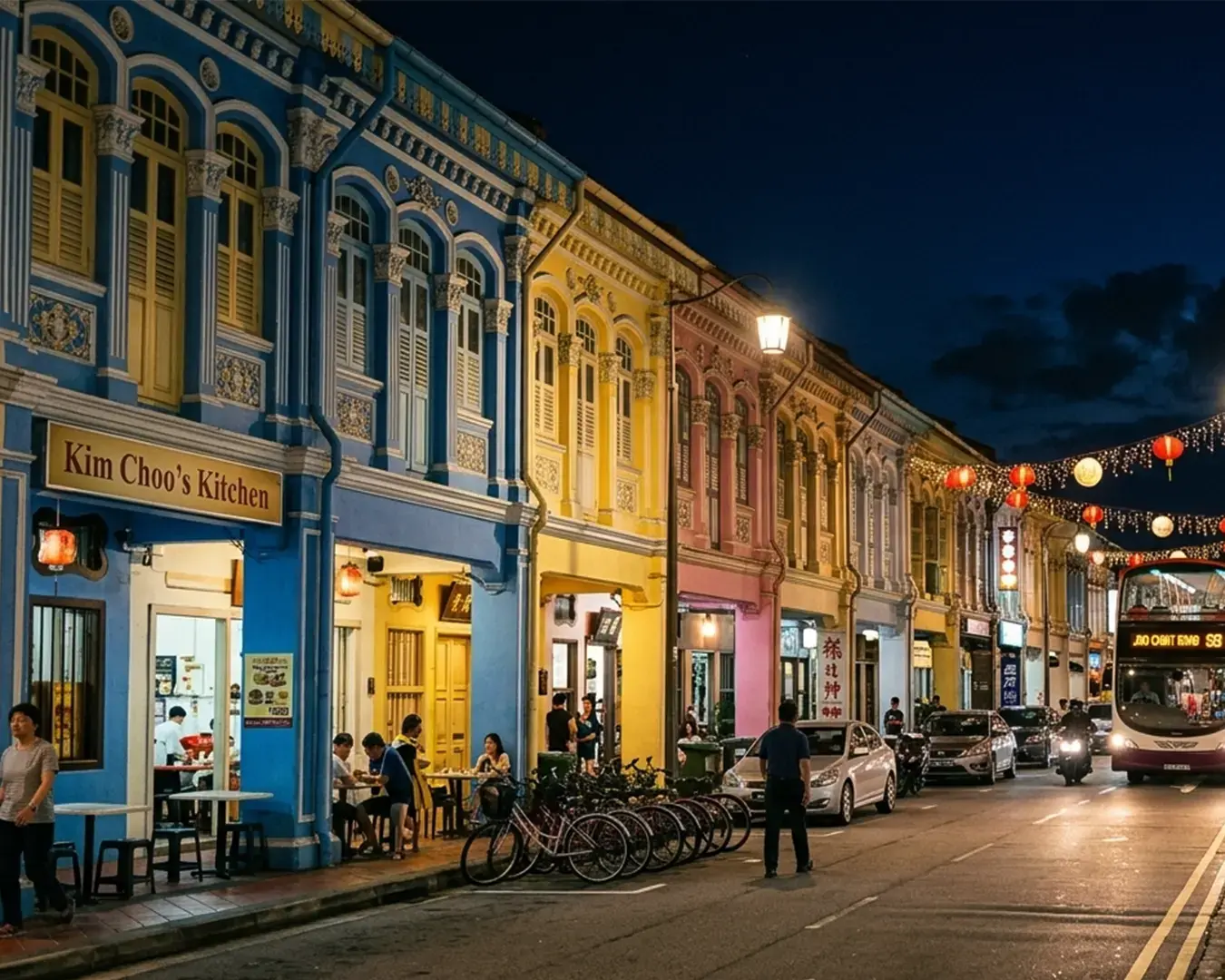 Colorful row of Peranakan shophouses at night, featuring blue, yellow, and pink facades. People dine outside, and a bus drives past under lanterns.