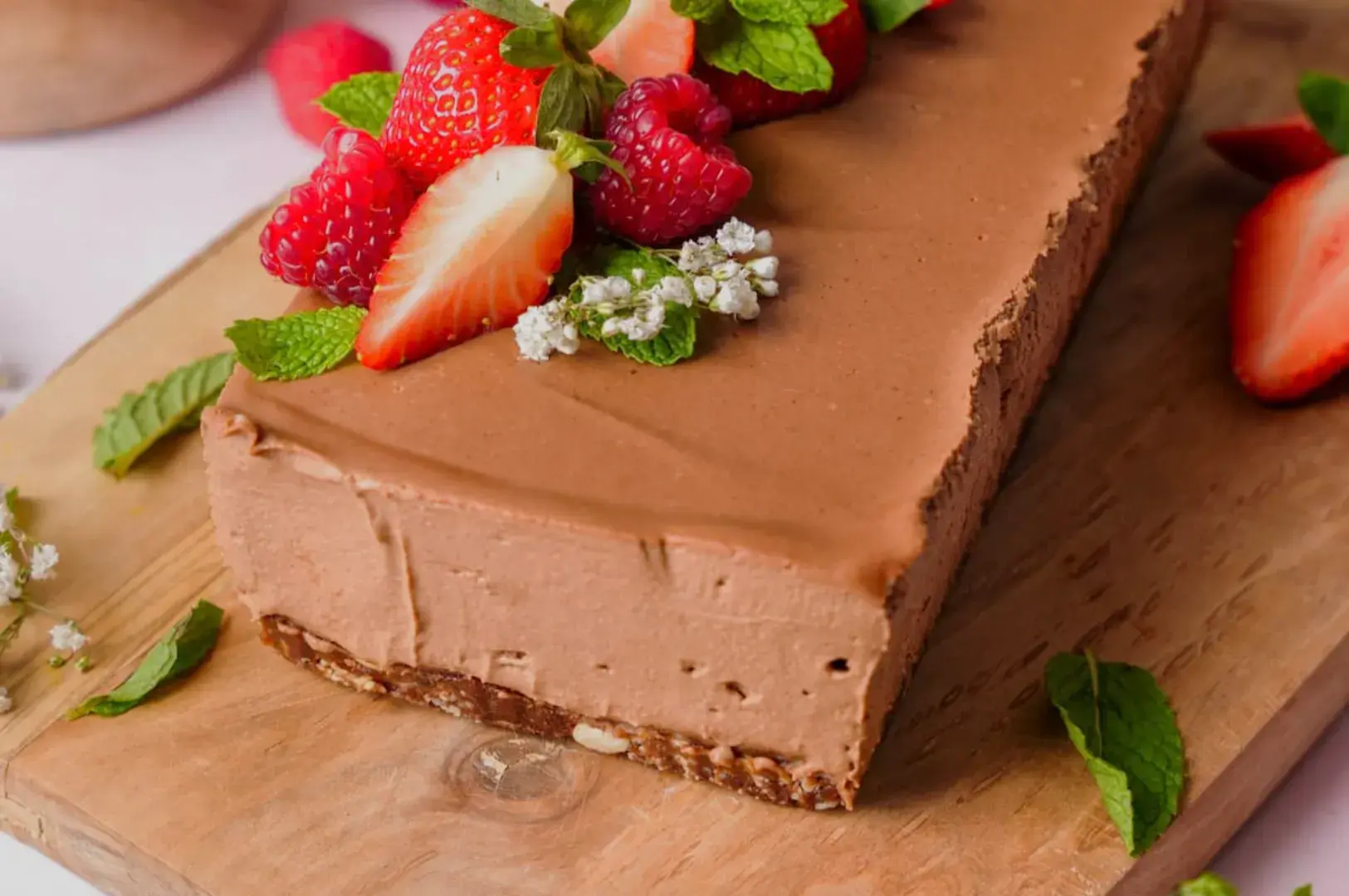 A chocolate cheesecake topped with fresh strawberries, raspberries, and mint leaves, presented on a wooden cutting board.