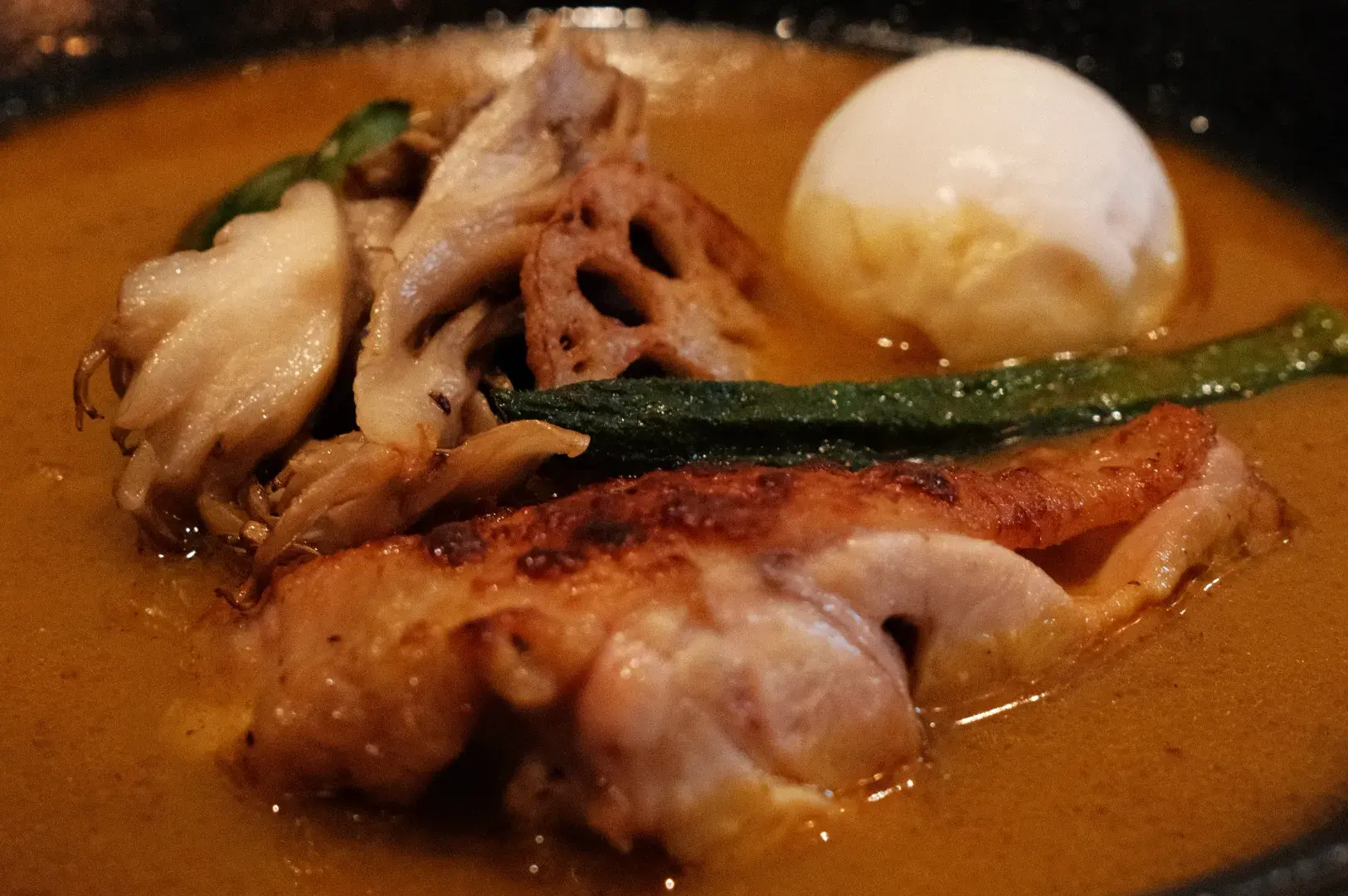 A bowl of soup curry featuring chicken and various vegetables, including mushrooms and green beans, with a poached egg on top.