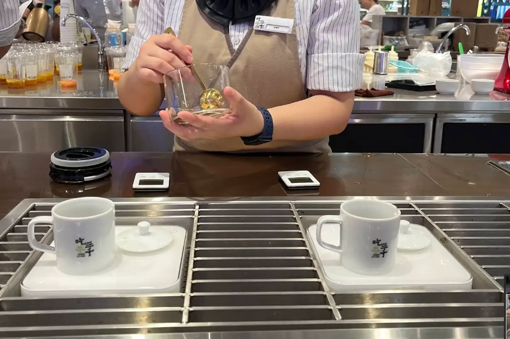 A CHICHA San Chen staff member in a striped shirt and beige apron meticulously measures ingredients with a spoon into a glass bowl over a polished dark countertop, situated behind two white teacups set on espresso-style extraction trays.
