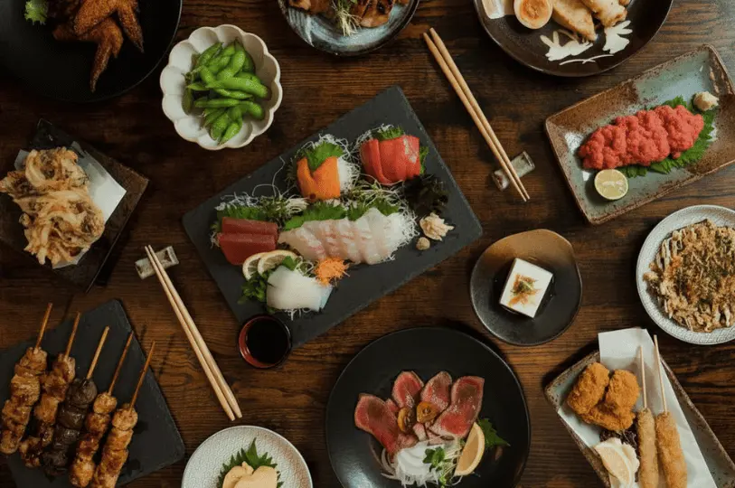 An overhead flat-lay view of a dark wooden table covered in a rich spread of Japanese dishes, featuring fresh sashimi, grilled yakitori skewers, edamame, and assorted fried foods.