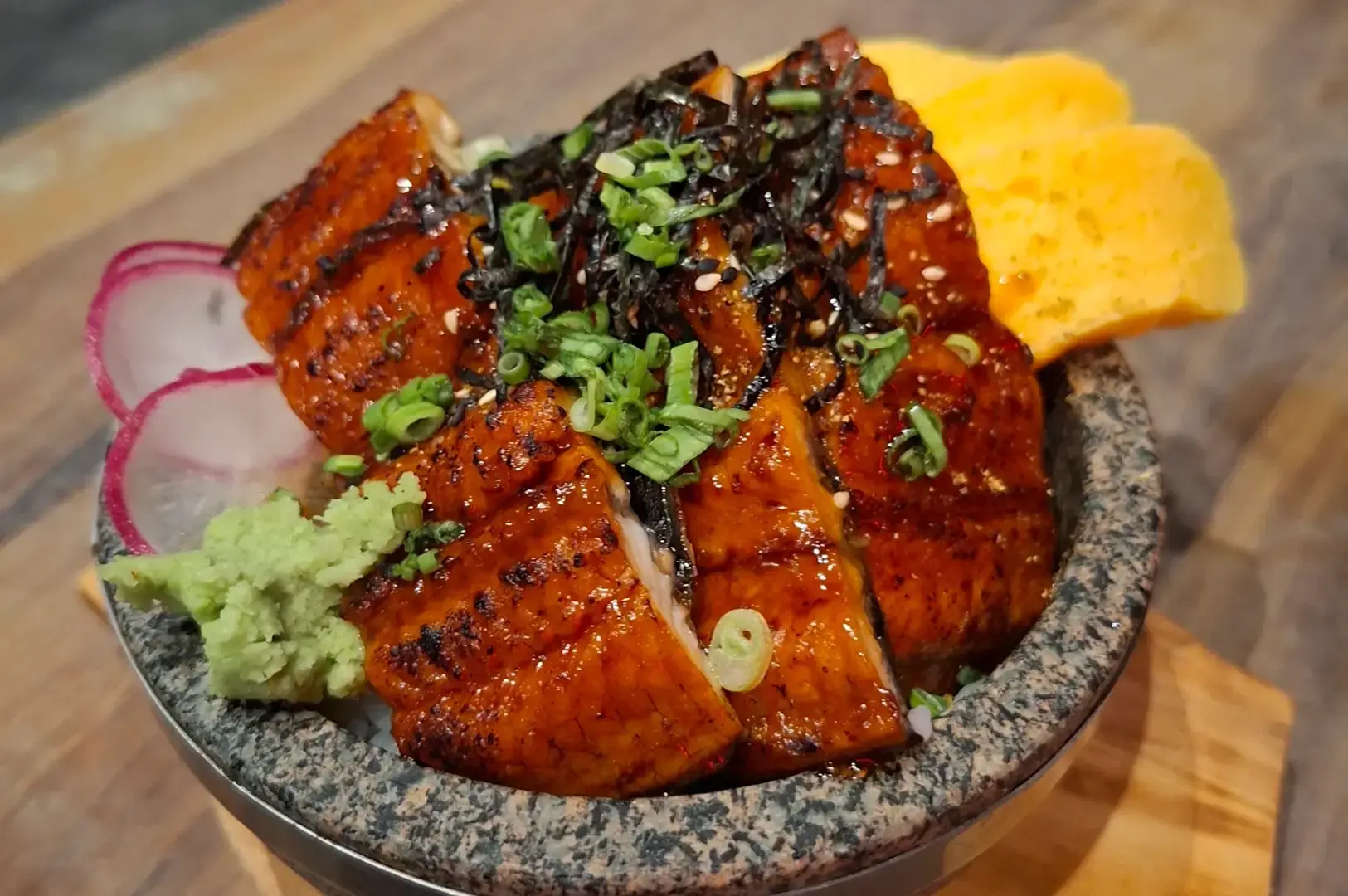 Glazed grilled eel fillets topped with chopped green onions and seaweed, served with wasabi, pickled radish, and rolled egg on a stone plate.