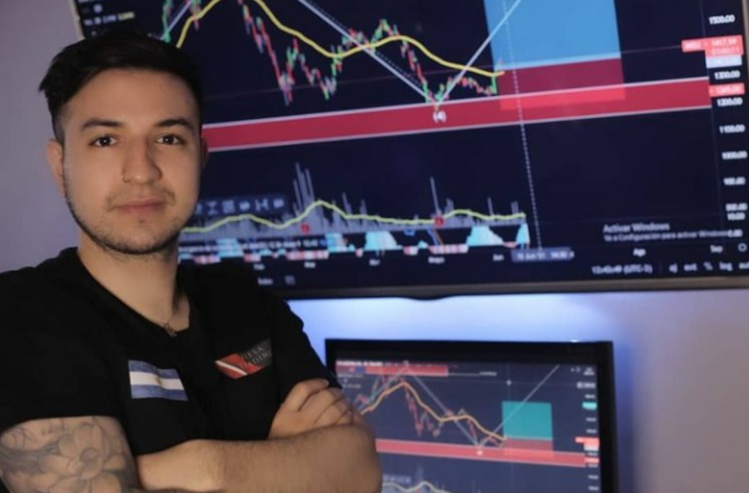 A young male trader with a sleeve tattoo sits with arms crossed in front of a multi-screen trading setup, wearing a branded black t-shirt. Behind him, a large monitor displays a detailed candlestick chart with key support and resistance zones marked in red, a moving average overlay, and volume indicators — conveying the image of a serious, experienced retail trader in his element.