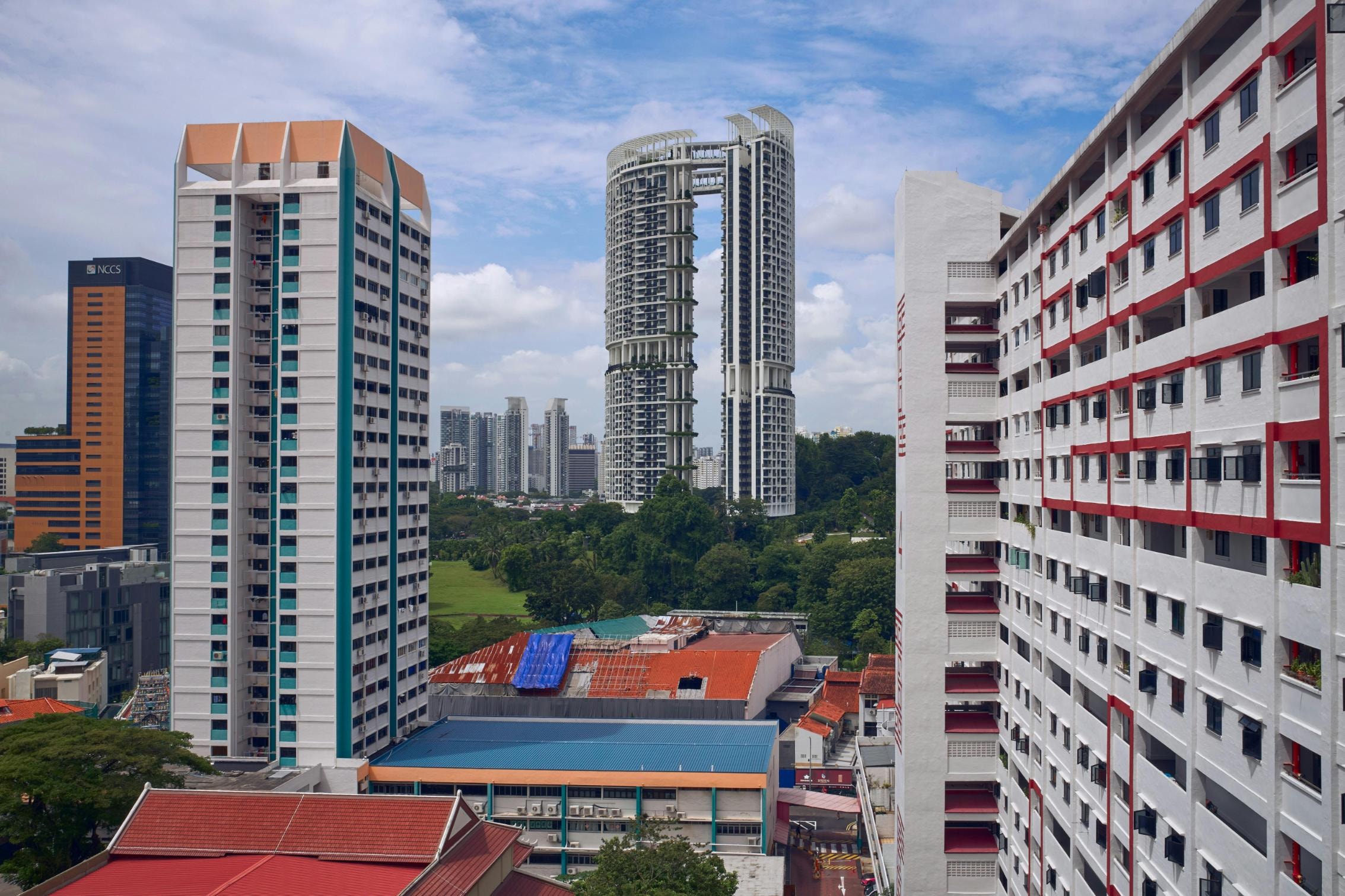 Tall, modern apartment complexes frame a distant view of a distinctive skyscraper featuring twin towers connected by a skybridge. The urban landscape is punctuated by buildings with white facades accented in red and teal, overlooking a patch of green trees under a partly cloudy sky.