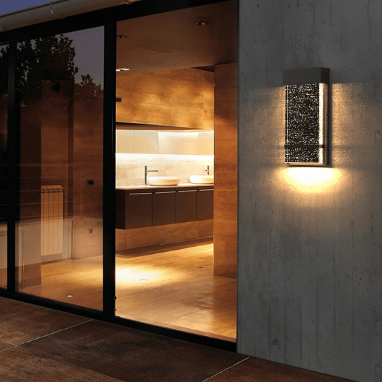 Modern wall-mounted outdoor light illuminating a concrete exterior next to large glass doors, revealing a warmly lit contemporary kitchen inside.