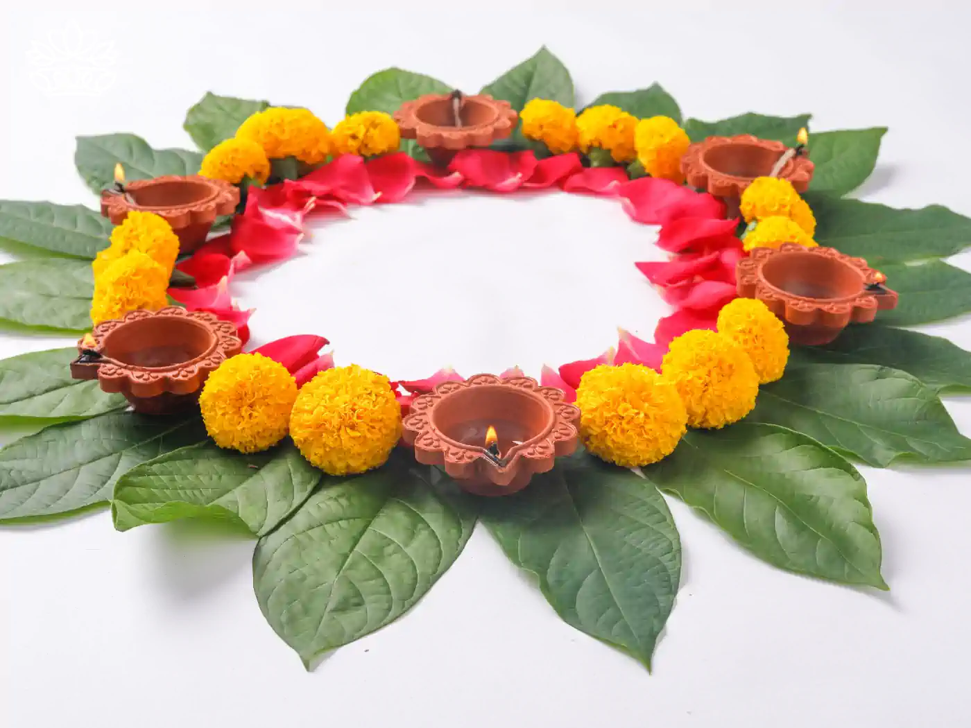The Festival marks, A circular arrangement of bright yellow marigolds and red rose petals on green leaves, interspersed with lit clay oil lamps, creating a festive and traditional Diwali decoration. Fabulous Flowers and Gifts - Diwali Flowers. Delivered with Heart.