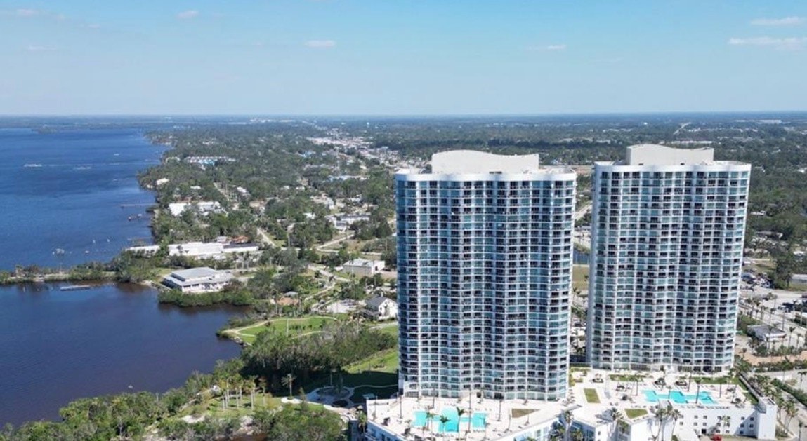 An aerial view showcases twin luxury towers of Oasis Condominiums rising majestically above the Caloosahatchee River, with the vibrant city skyline of downtown Fort Myers in the background, offering residents glorious waterfront views and a perfect setting for enjoying Florida sunsets. These luxury waterfront residences feature private balconies, allowing for unobstructed panoramic views of the scenic surroundings.
