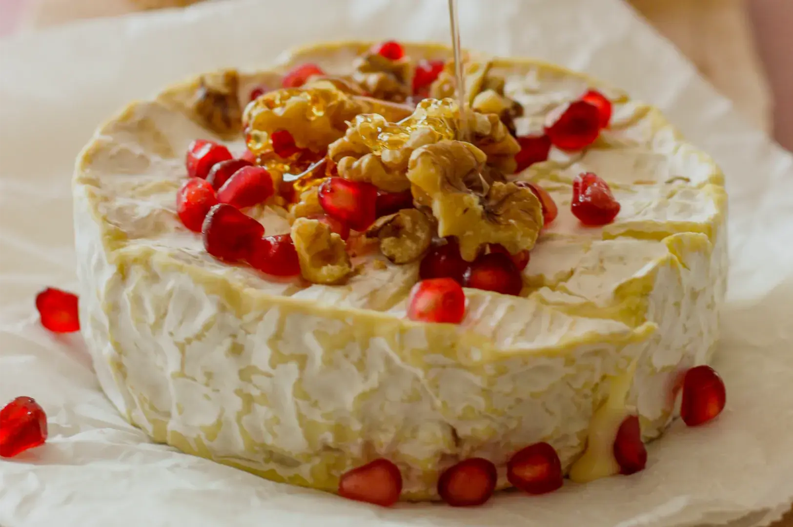 A creamy round cheese wheel topped with walnuts, bright red pomegranate seeds, and a drizzle of golden honey, presented on parchment for an inviting appetizer.