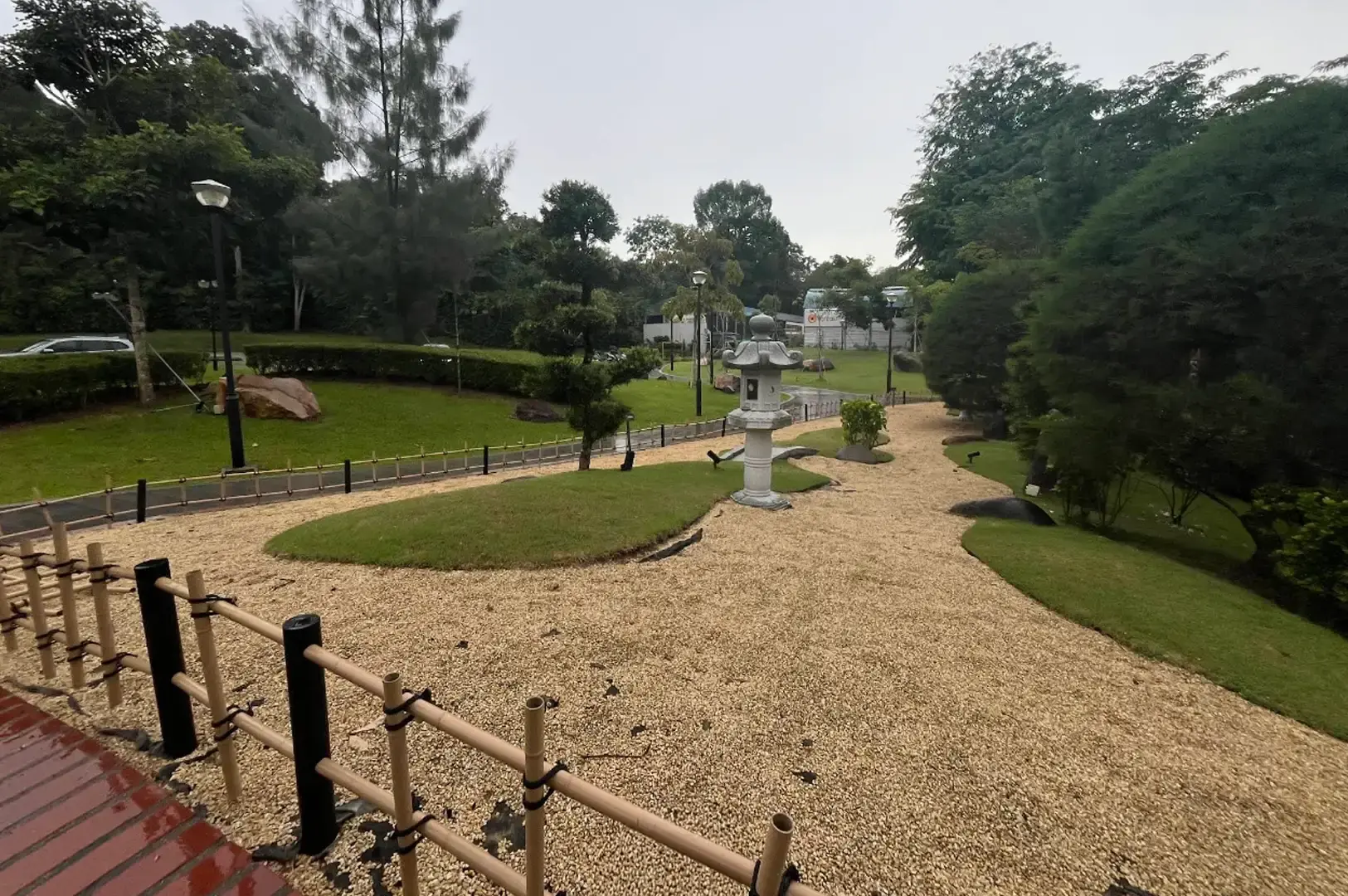 A Japanese zen garden area featuring neatly raked light brown gravel, a traditional stone lantern (tōrō), grass mounds, and a low bamboo fence running alongside a red-tiled path under overcast skies.