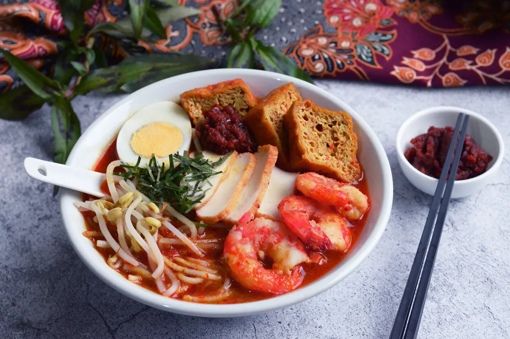 A vibrant bowl of spicy laksa is garnished with prawns, boiled egg, tofu, bean sprouts, and herbs. Beside it, a small bowl of chili paste and chopsticks add an inviting touch.