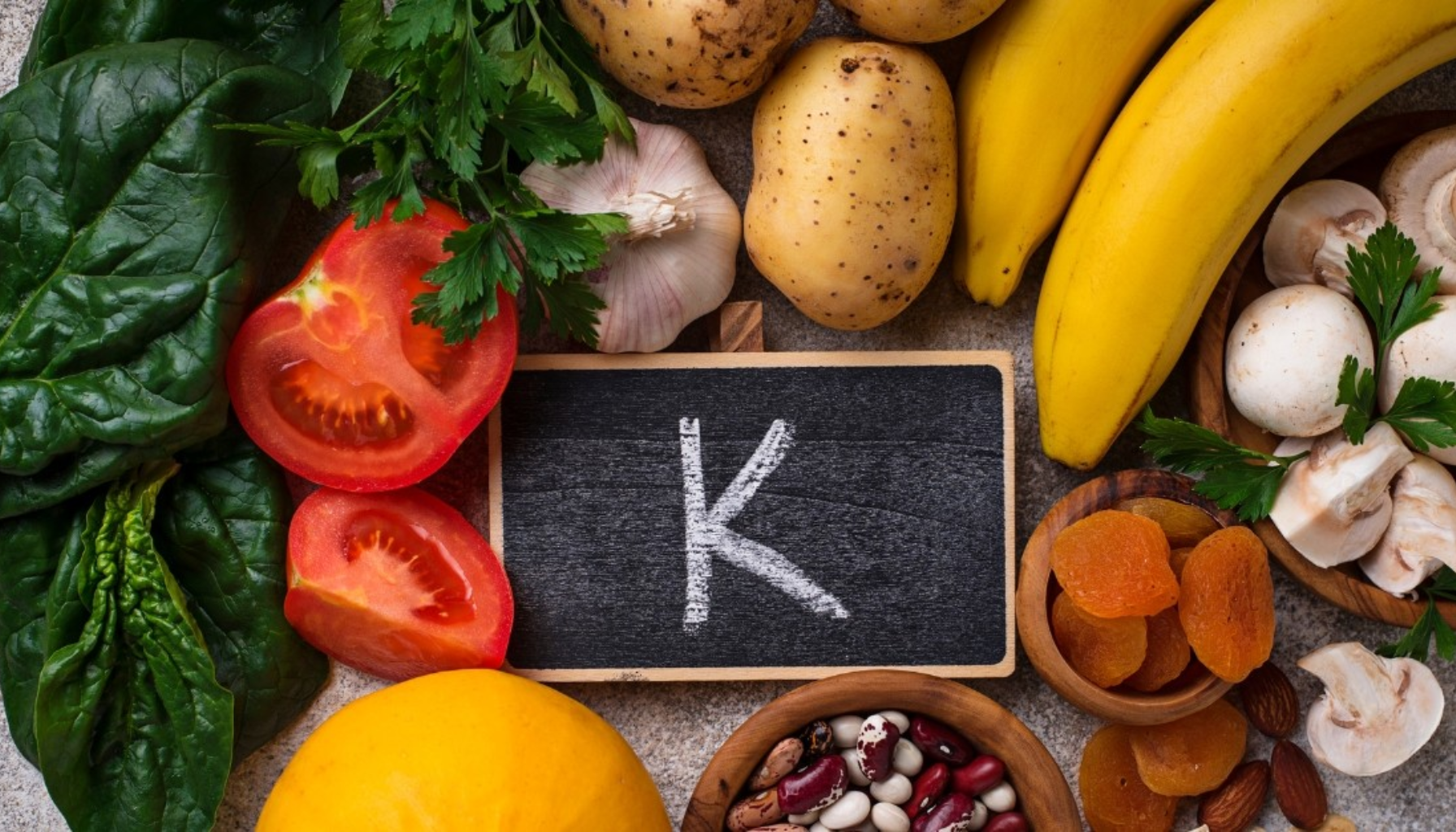 "Ulike matvarer rike på kalium som bananer, spinat, tomater og poteter, med en tavle som viser bokstaven K i midten."