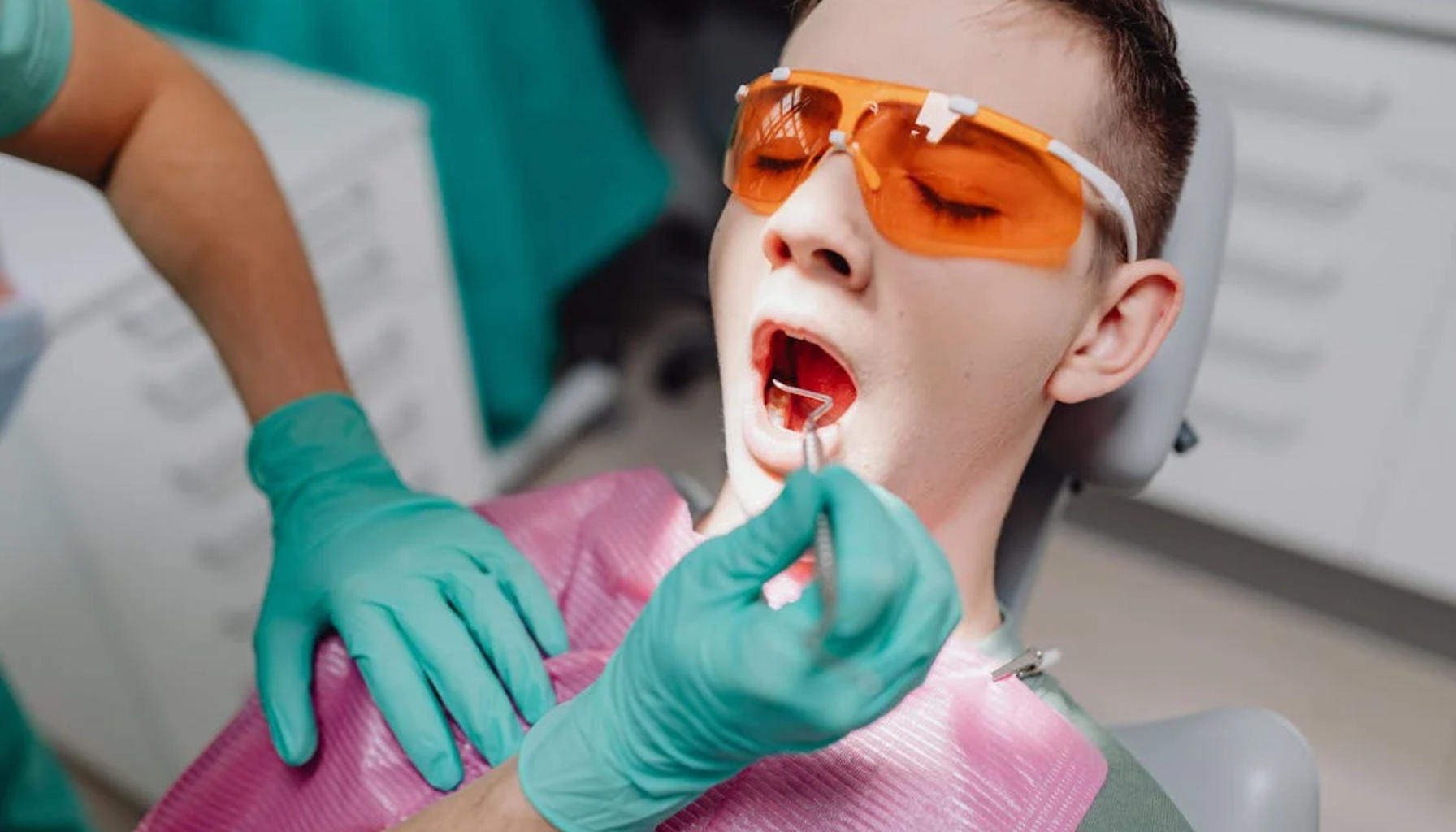 Ung mann med oransje beskyttelsesbriller ligger i tannlegestol og får munnen undersøkt. Tannlegen bruker grønne hansker og tannspeil. Moderne klinikkmiljø.