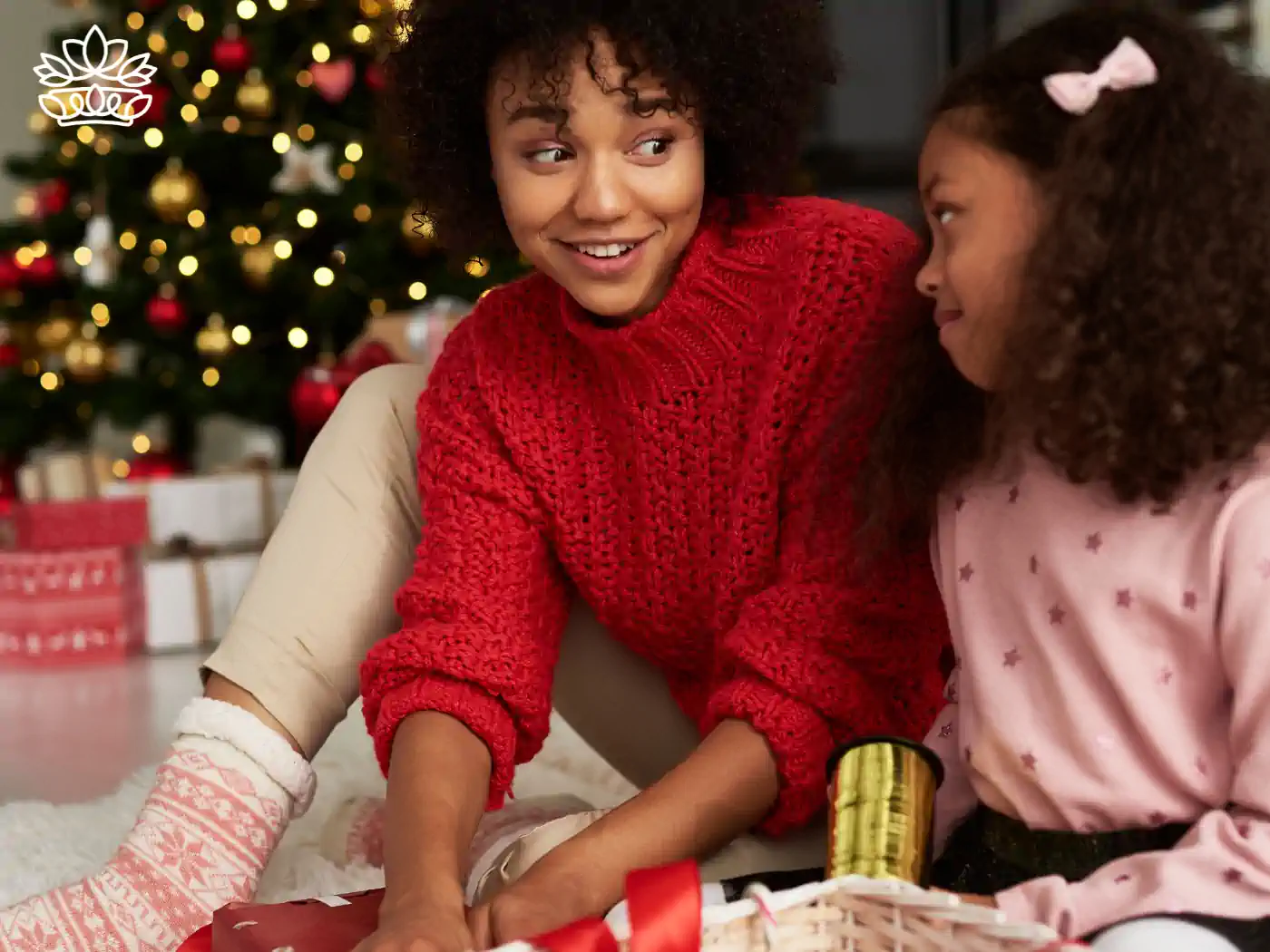 A mother and child wrapping presents together, surrounded by gifts and twinkling lights near a Christmas tree, part of the Festive Season Gift Boxes Collection. Delivered with Heart by Fabulous Flowers and Gifts.