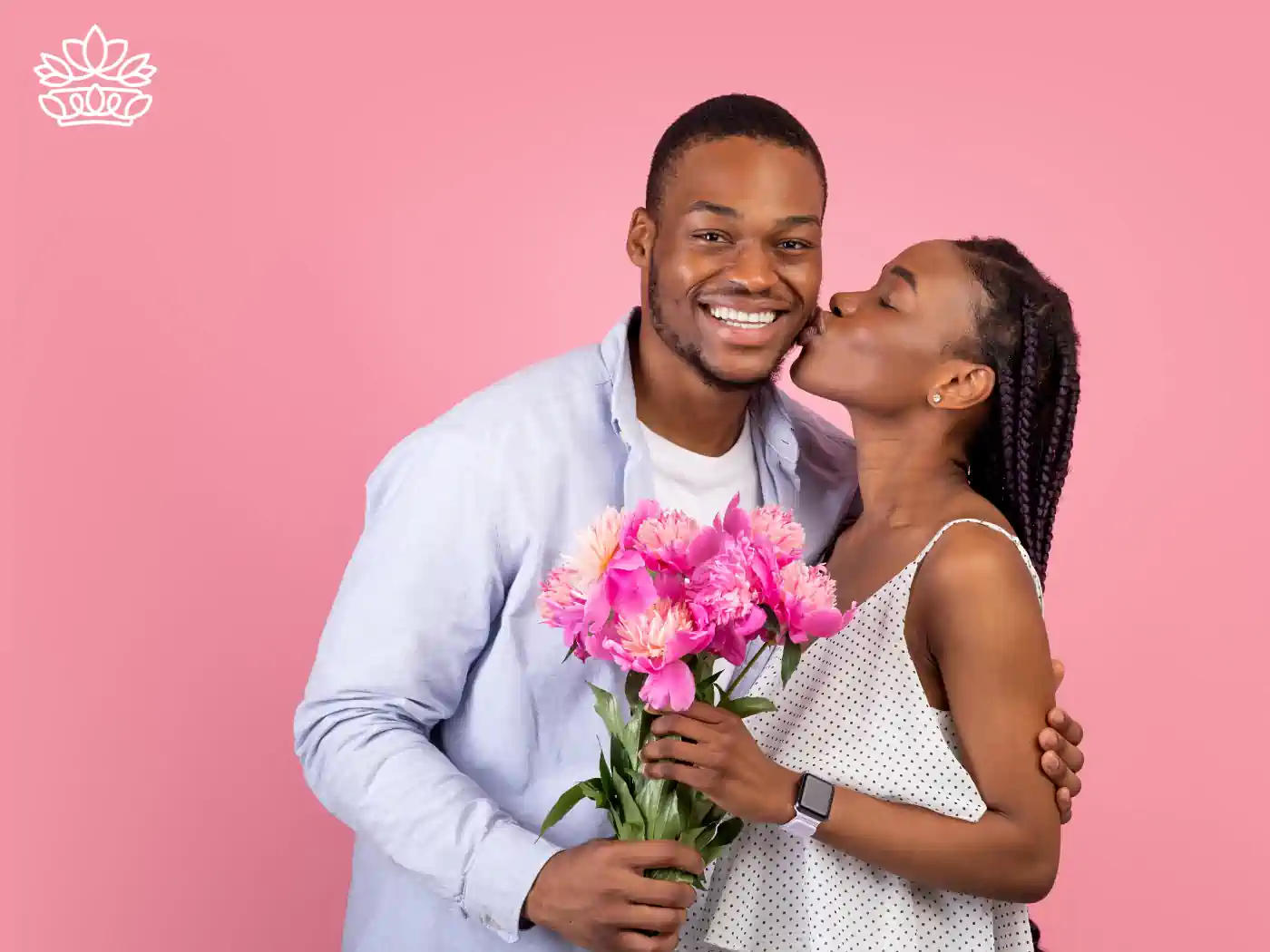 A woman kisses her partner on the cheek as he smiles and holds a bouquet of pink peonies, set against a soft pink background. Fabulous Flowers and Gifts delivered with heart. Valentine's Day Flowers.