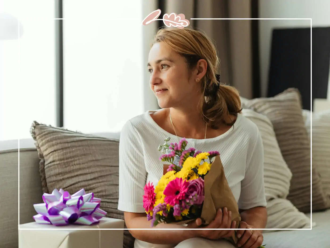 A woman holding a small bouquet of colorful flowers, sitting next to a gift with a purple ribbon. Fabulous Flowers and Gifts.