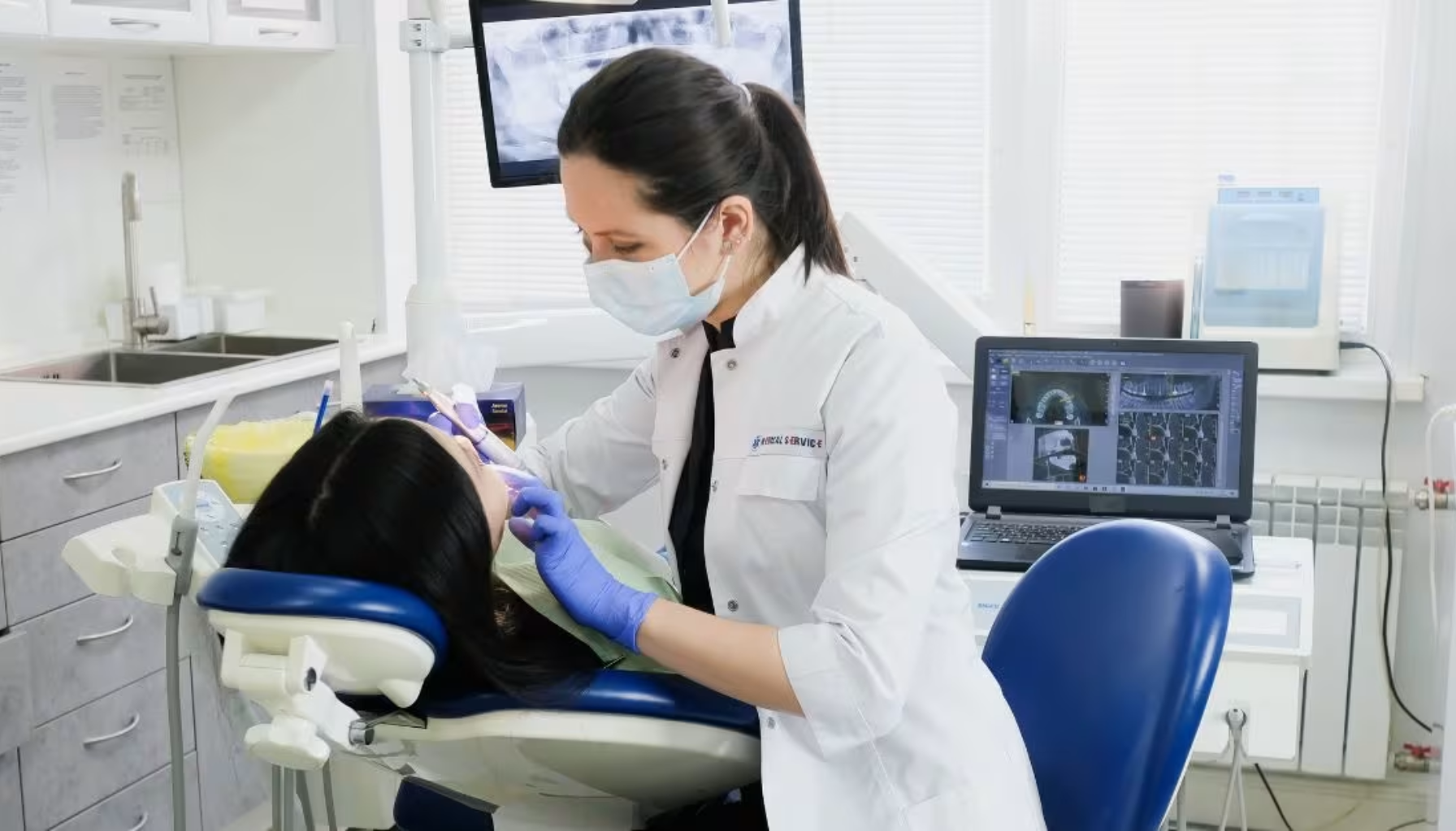 Tannlege i arbeid med pasient i tannlegestol, med dataskjerm som viser tannrøntgen.
