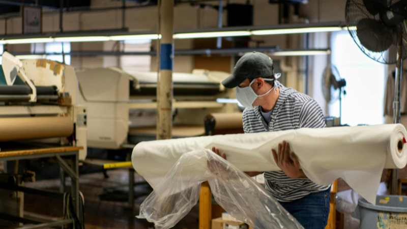 A person wearing a mask and cap carries a large roll of white fabric in a factory setting.