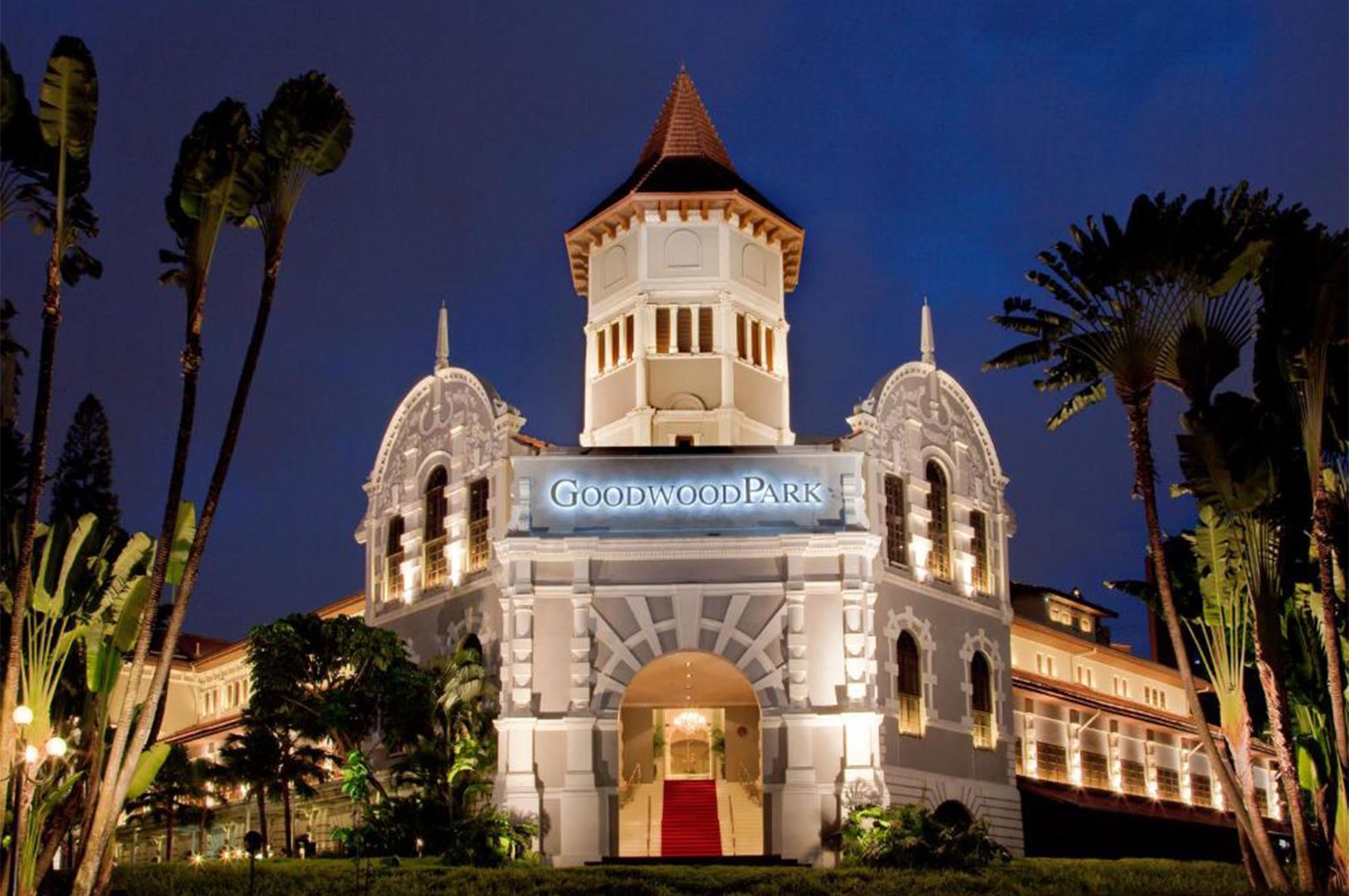 Night view of Goodwood Park Hotel, a historic white building with a central tower and illuminated façade surrounded by palm trees.