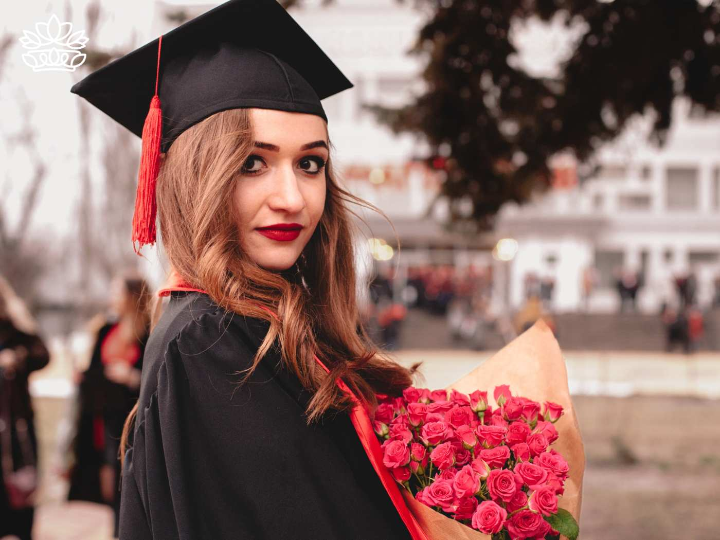 Confident young woman in graduation cap and gown holding a bouquet of red roses, looking back with a thoughtful expression. Graduation Flowers Delivered with Heart by Fabulous Flowers and Gifts.