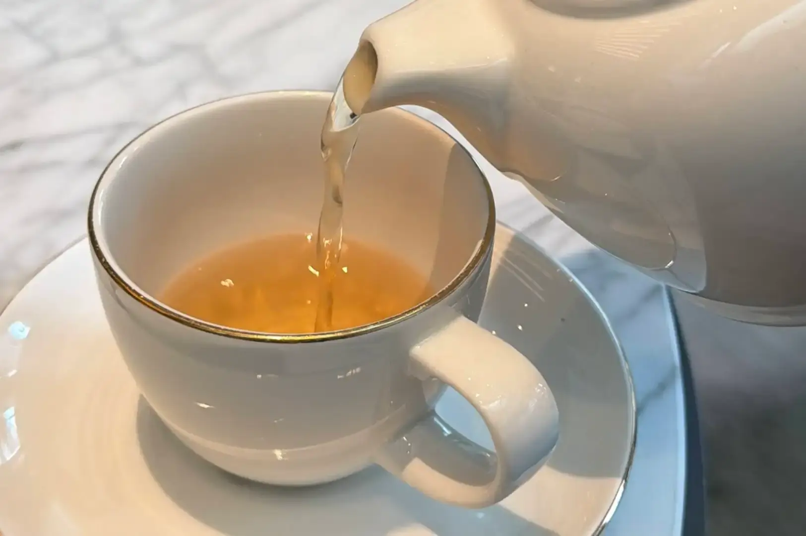 A close-up shot captures clear hot tea being poured from the spout of a white ceramic teapot into a white teacup with a thin gold rim, sitting on its matching saucer over a light marble surface.