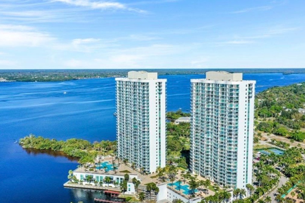 The image depicts modern luxury towers with sleek glass facades and private terraces that offer glorious waterfront views of the Caloosahatchee River, embodying the essence of luxury waterfront residences in Fort Myers. Residents can enjoy relaxing evenings while admiring the vibrant city skyline and famous Florida sunsets from their own units.