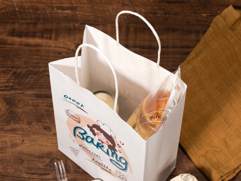 White bakery paper bags with sturdy handles. Elegant and stylish print makes them an attractive option for brand promotion. Filled with fresh bread and packaged treats, the bag highlights eco-friendly convenience while promoting brand identity. These bags are ideal for bakeries, cafés, and food shops offering quality goods to customers. 