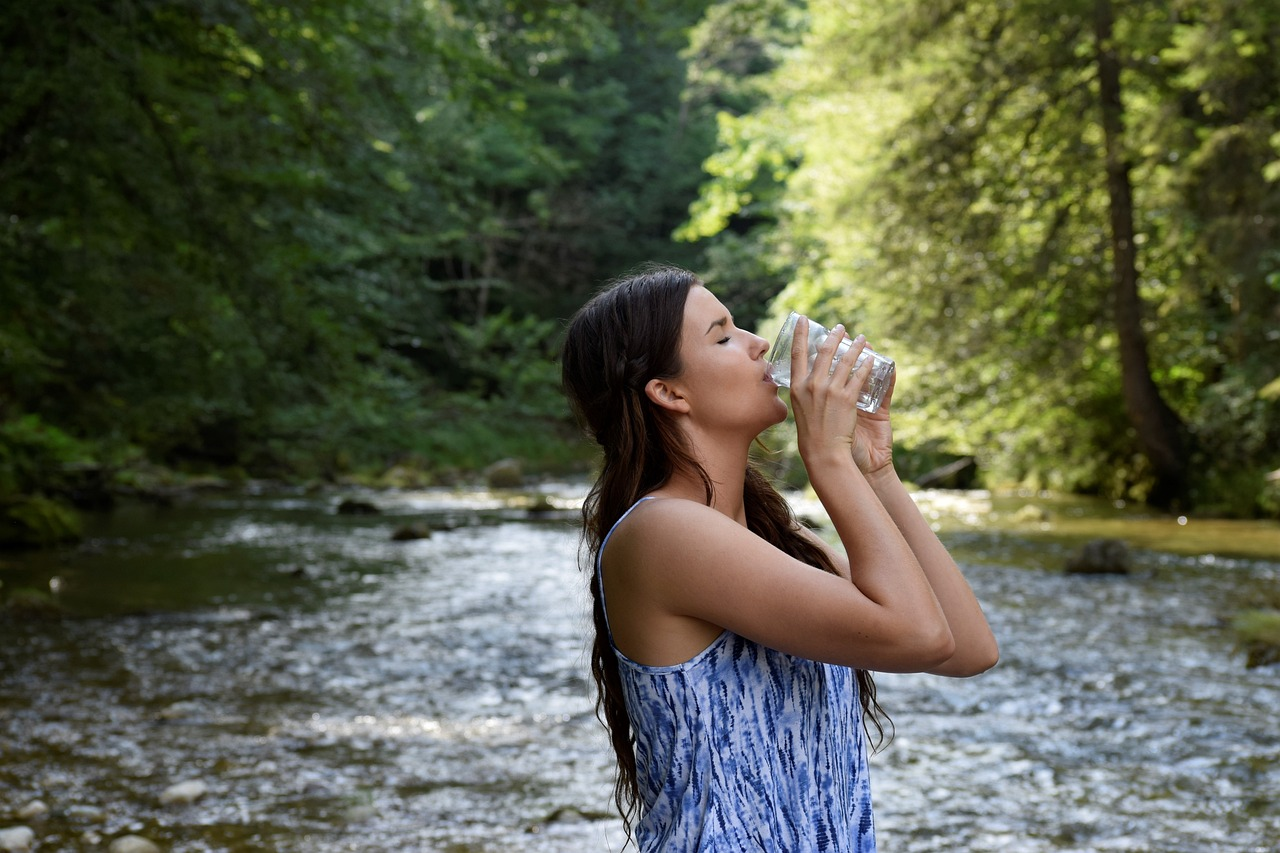 An image of a young woman drinking a glass of water in front of a lake.