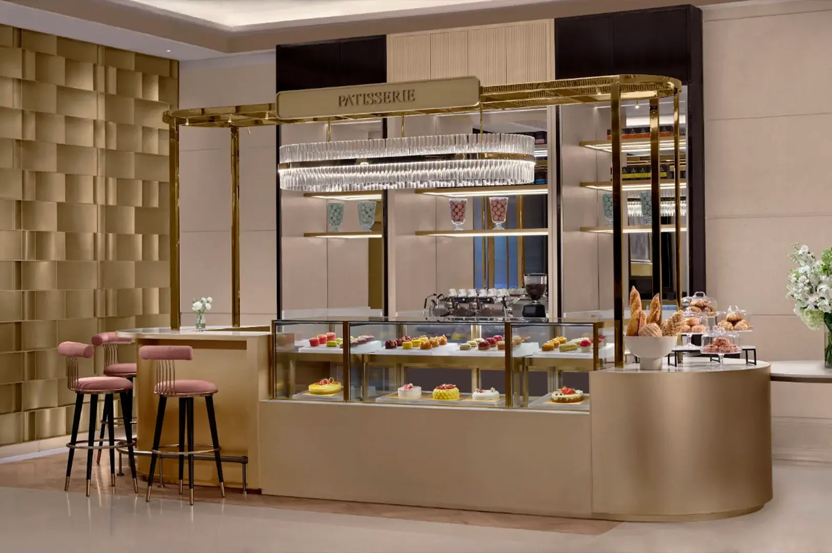 This eye-level shot captures a luxurious, modern patisserie counter inside a hotel, featuring a sleek curved structure finished in a brushed gold metallic sheen. The glass display case is filled with an array of colorful miniature cakes, tarts, and whole pastries, illuminated by an ornate, multi-layered crystal oval chandelier hanging from a gold frame labeled "PATISSERIE". To the left, three high-top bar stools with pink velvet cushions and slender black legs with gold accents line the counter, while a selection of fresh baguettes, croissants, and baked goods are displayed on stands to the right next to a vase of fresh white flowers against a textured gold block wall.