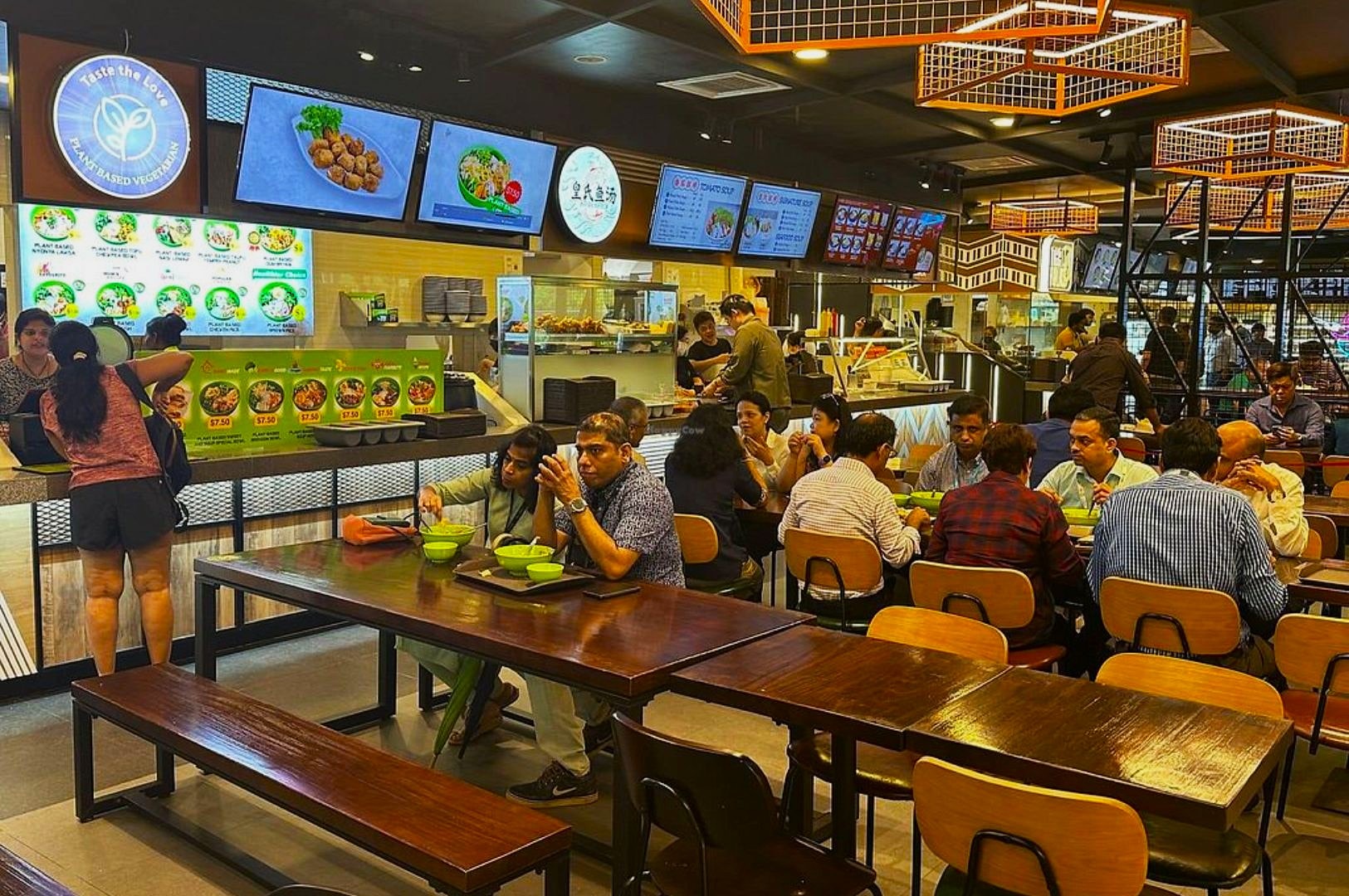 A bustling food court with people dining at wooden tables; digital menus display various Asian dishes. The atmosphere is lively and vibrant.