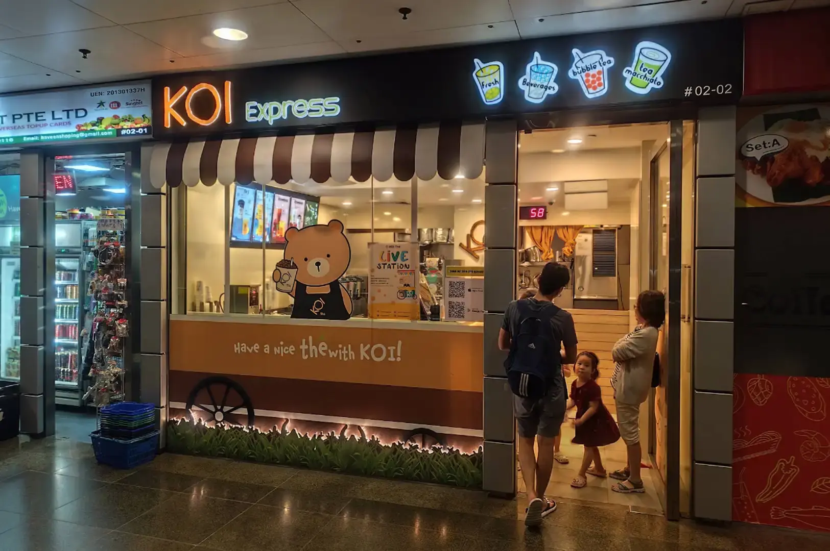 The exterior of a KOI Express shop styled like a vintage food truck, complete with a brown-and-white awning, a cute cartoon bear decal holding a boba cup in the window, glowing drink menu icons along the top header, and customers entering the store.