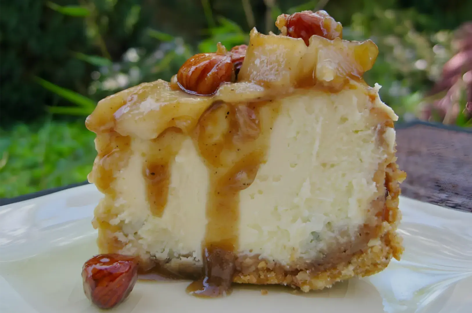 A creamy slice of cheesecake topped with caramel sauce and glazed nuts sits on a white plate outdoors, creating a rich, tempting dessert scene.