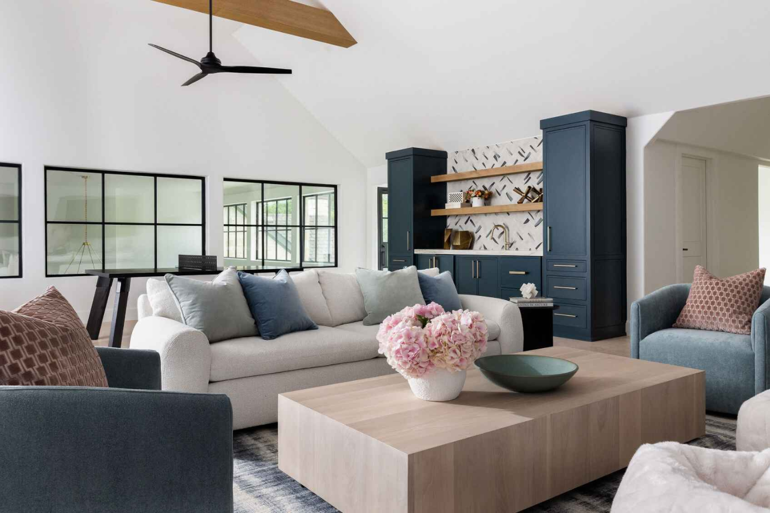 Bright living space with vaulted ceilings, white sofa and blue accent chairs, light wood coffee table, and a navy built-in bar with open shelving.