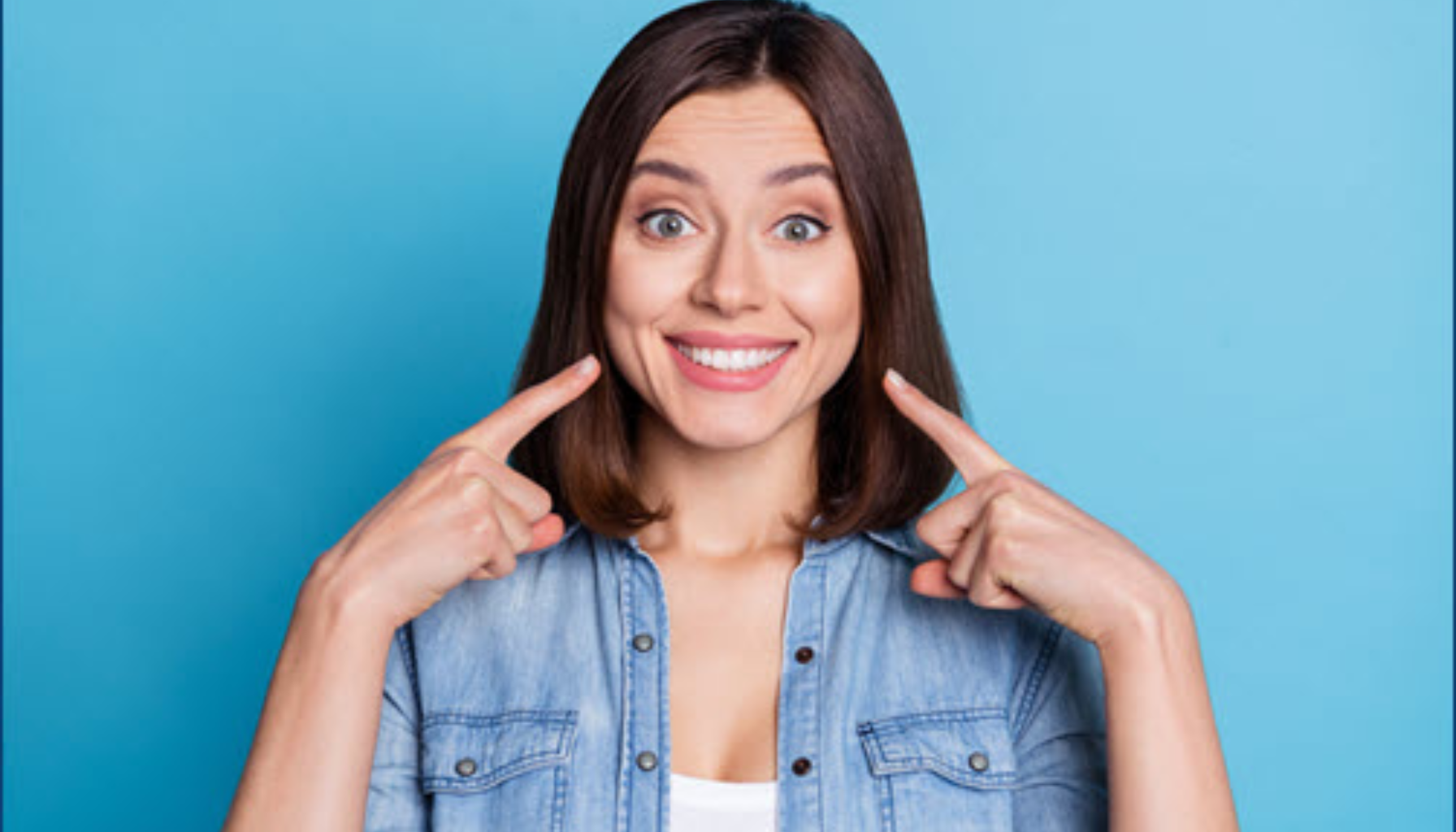 Kvinne peker på sitt brede smil og viser hvite tenner foran blå bakgrunn.