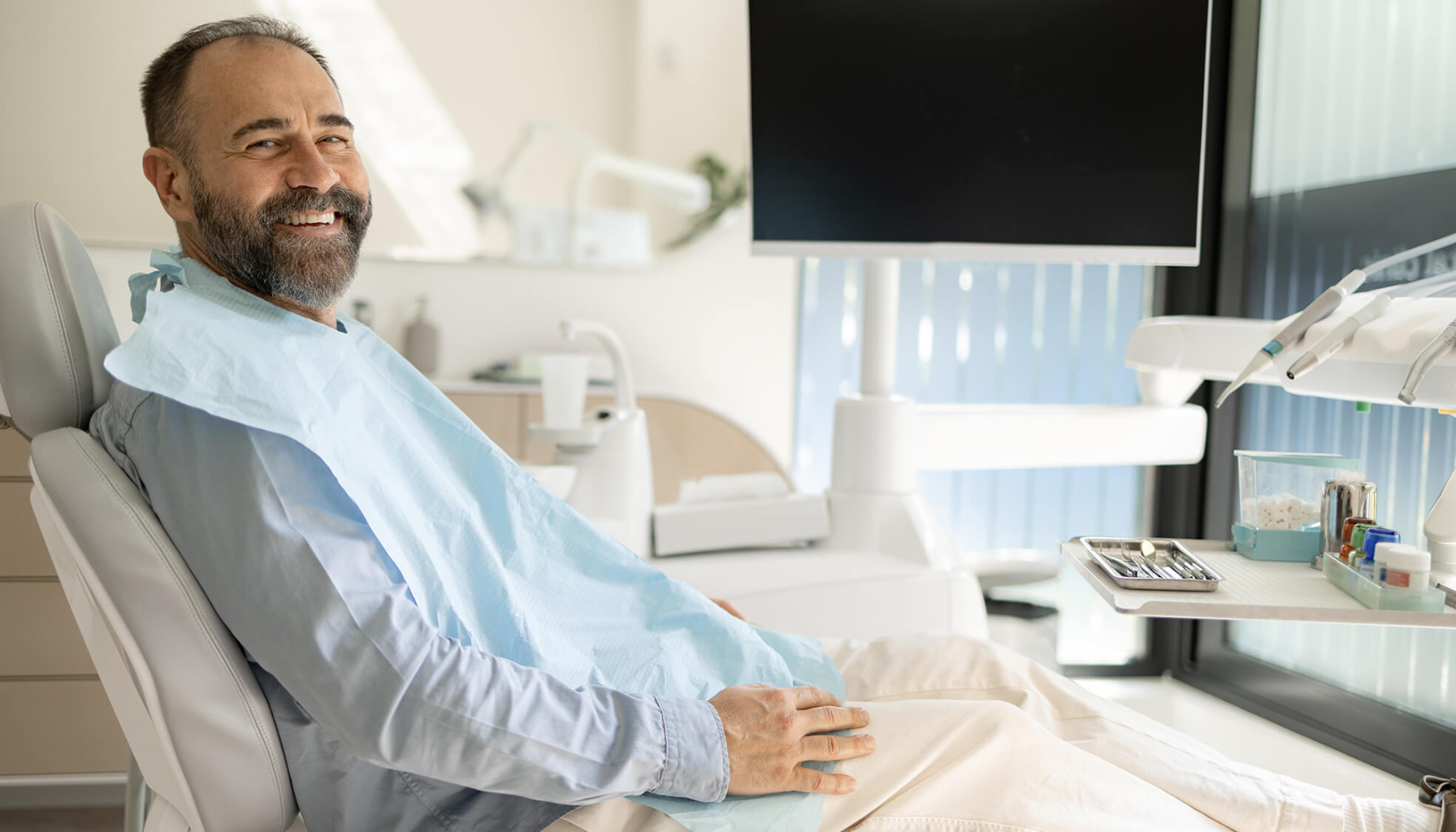 Refusjon hos tannlege: Hva du må vite før du betaler 12 Smilende mann sitter avslappet i tannlegestol med beskyttelsesforkle, omgitt av moderne utstyr i lys klinikkmiljø.