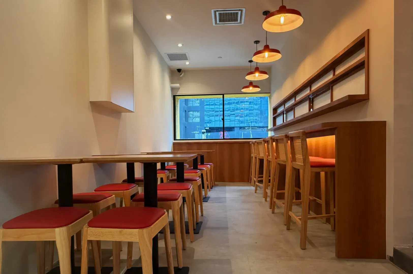 A modern, minimalist interior of a restaurant featuring wooden tables and chairs with red cushions, warm lighting, and large windows.